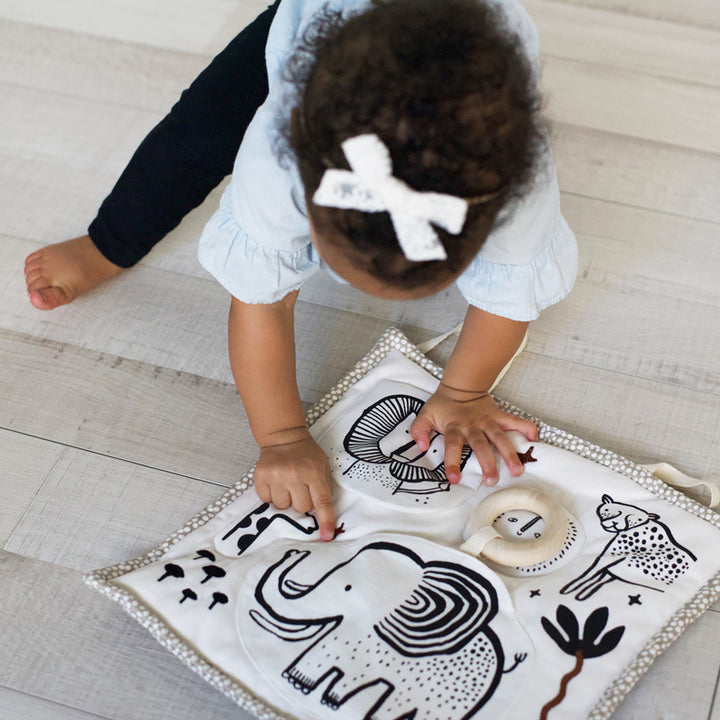 A toddler girl is touching an embroidered star on a safari themed organic activity pad with elephant, lion, cheetah, zebra, and sun.