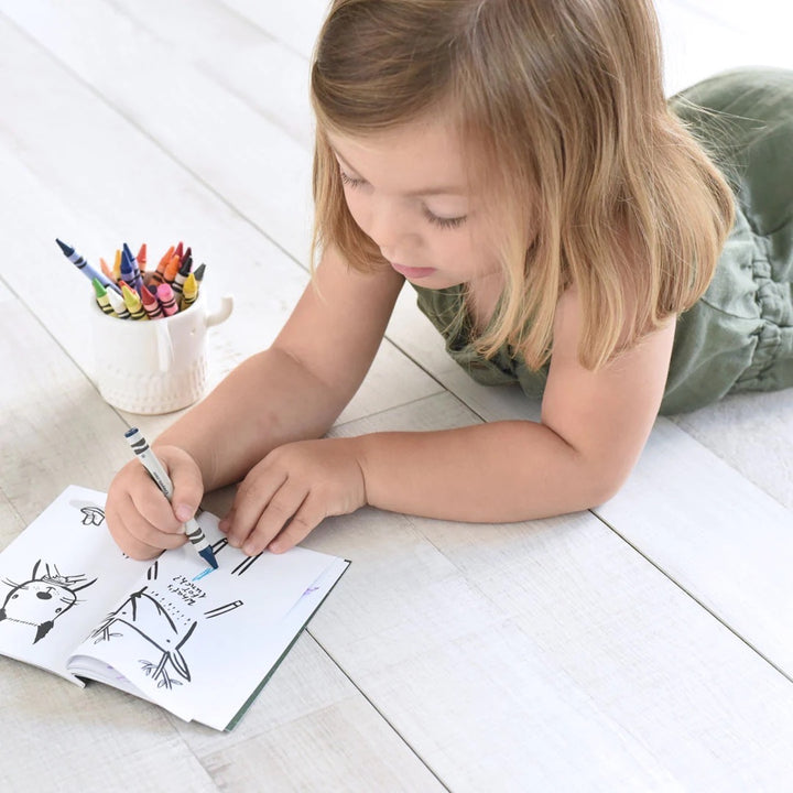 Young child laying on the floor and coloring with crayons in a small book of illustrated animals.