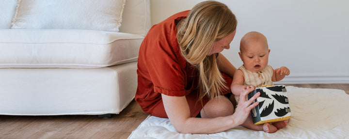 Mother sitting on the floor with her baby who is engaged with a black and white soft activity block.