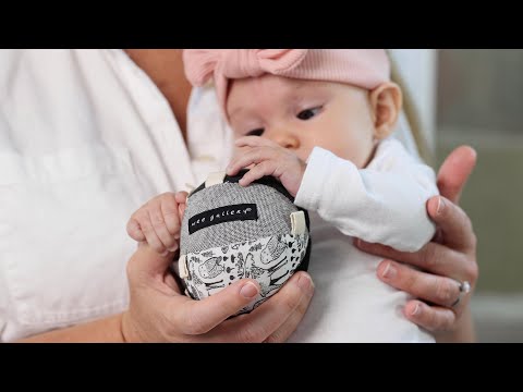 Video shows a 6 month old baby playing with a Wee Gallery Taggy Ball. She grips it and pulls the tags while sitting in her mom's lap. She turns the ball, gazes at it, and smiles.